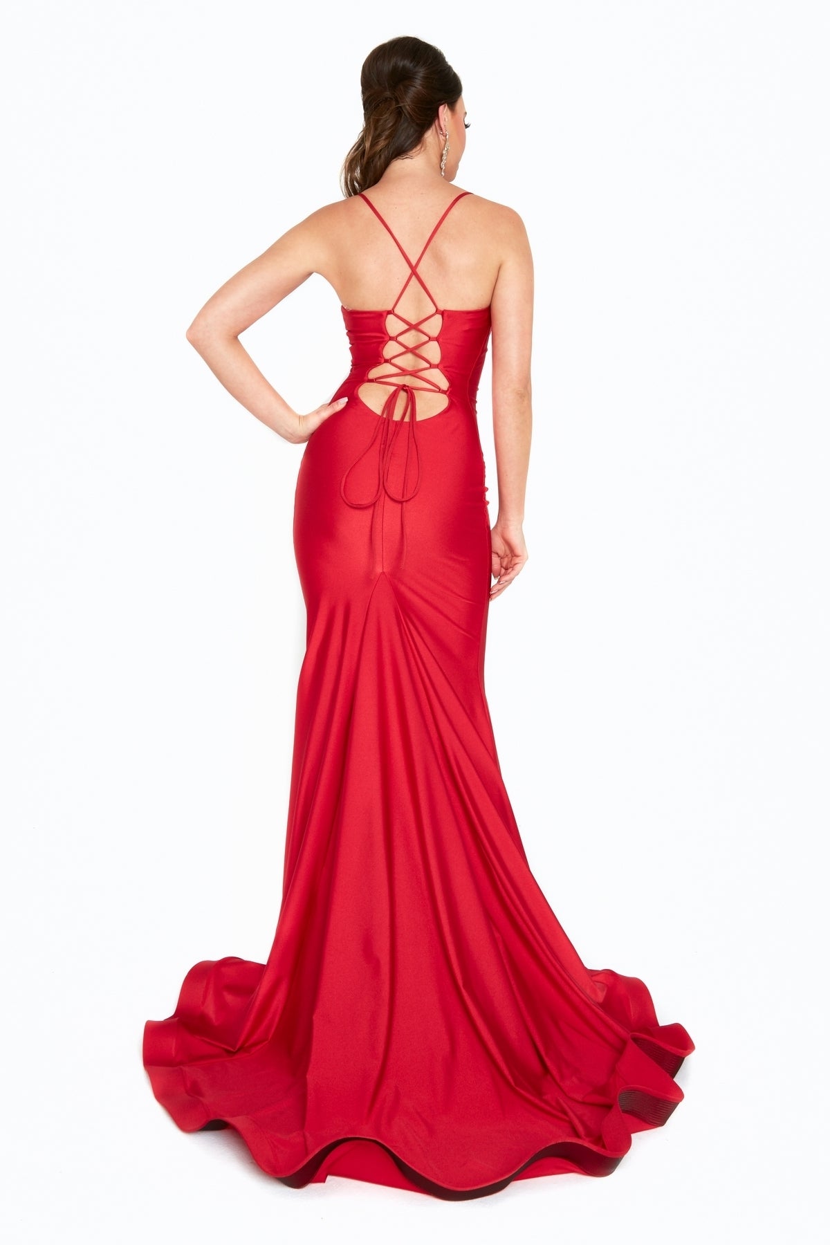 A woman in the Lace-Up Back Long Prom Dress: Atria 6520H, featuring a fitted red mermaid silhouette with an open lace-up back and crisscross straps, stands facing away from the camera.